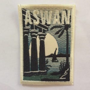 Puede incluir: Parche bordado con la palabra "ASWAN". El diseño muestra columnas, una palmera, un sol y agua, en tonos negros, azules y verdes. Tiene un borde color crema, ideal para coser en ropa o bolsos.