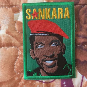Może przedstawiać: Haftowana naszywka przedstawiająca Thomasa Sankarę, z napisem "SANKARA" w żółtych literach powyżej. Portret przedstawia Sankarę w czerwonym berecie. Naszywka ma zieloną obwódkę i jest umieszczona na wzorzystym tle.