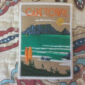 May include: Embroidered patch depicting Cape Town scenery. The design shows the text "CAPE TOWN" above a mountain range, ocean, and two orange surfboards. The patch has a white border and is set against a patterned background.