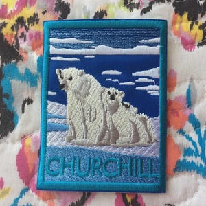 Op de afbeelding: Geborduurde patch met een ijsberen familie op een blauw-witte achtergrond, met het woord "CHURCHILL" in teal. De patch heeft een teal rand en toont een besneeuwd landschap met wolken.