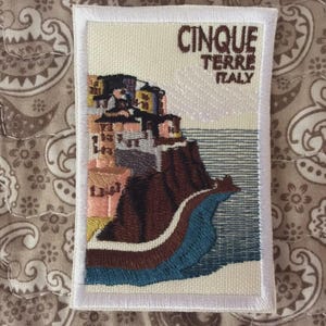 Può includere: Toppa ricamata raffigurante un paesaggio costiero delle Cinque Terre, Italia. La toppa raffigura edifici colorati su una scogliera, con la scritta "CINQUE TERRE ITALY" in marrone. La toppa ha un bordo bianco ed è rettangolare.