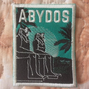 May include: Embroidered patch with the word "ABYDOS" in white block letters above a teal background. The patch features a black and white illustration of two seated figures and palm trees.