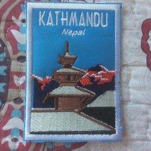 Może przedstawiać: Haftowana naszywka z napisem "KATHMANDU Nepal" w kolorze białym na niebieskim tle. Naszywka przedstawia budynek w stylu pagody i pasmo górskie. Naszywka ma białą obwódkę.