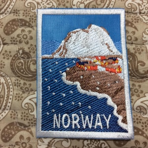 Peut inclure: Écusson brodé représentant un paysage côtier norvégien. Le motif comprend une montagne enneigée, une mer bleue et de petits bâtiments. Le mot "NORWAY" est brodé en blanc en bas.