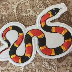 May include: Embroidered snake patch featuring a black, red, and yellow coral snake design. The patch has a white border and is set against a paisley patterned background. This patch is ideal for jackets, bags, or other fabric items.