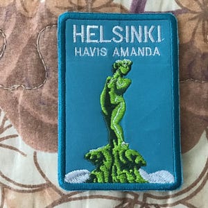 May include: A rectangular patch with a teal background and a teal border. The patch features the text "HELSINKI HAVIS AMANDA" in white. A green statue of a woman stands on a green base. The patch is likely a souvenir or collectible item.
