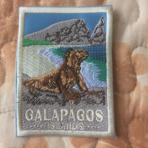 May include: Embroidered patch depicting the Galapagos Islands. It shows a brown iguana on a rocky shore, with a blue sea and grey rock formations. The text "GALAPAGOS ISLANDS" is embroidered at the bottom of the patch.