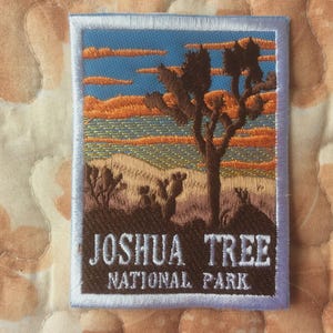 May include: Embroidered patch depicting a desert scene with a Joshua tree under a blue sky and orange clouds. The patch reads "JOSHUA TREE NATIONAL PARK" in white text on a brown base, framed by a light blue border.