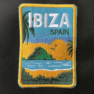 May include: Rectangular embroidered patch with the word "IBIZA" in white capital letters above "SPAIN". The design depicts a beach scene with palm trees, a green island, and a blue sea, set against a yellow and blue backdrop.
