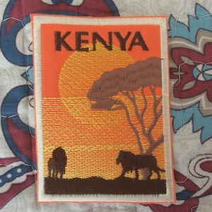 May include: Embroidered patch depicting a Kenyan sunset. The patch has an orange backdrop with a large sun, a tree, and two lions. The word "KENYA" is at the top in dark brown letters. The patch is rectangular with a white border.