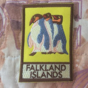 May include: Embroidered patch featuring three penguins with blue, white, and brown details on a yellow background. The patch has a brown border and the text "FALKLAND ISLANDS" in a matching brown color.