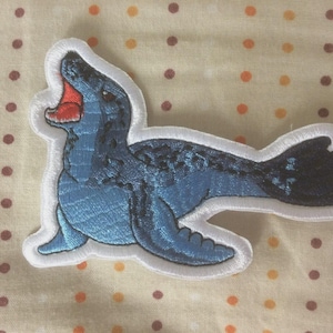 May include: Embroidered patch of a blue seal with a white border. The seal has a red mouth and black spots on its back. The patch is set against a beige background with orange and brown polka dots.