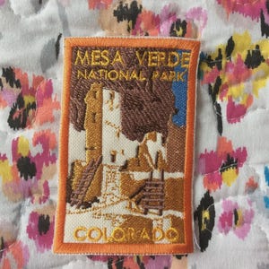 May include: Embroidered patch depicting Mesa Verde National Park in Colorado. The rectangular patch has an orange border and shows a detailed illustration of the park's structures in shades of brown, tan, and white, with the park name and state name.