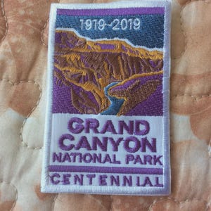 May include: Embroidered patch featuring a landscape of the Grand Canyon in shades of brown, purple, and blue. The patch includes the text "1919-2019" at the top and "Grand Canyon National Park Centennial" at the bottom, all in white lettering.