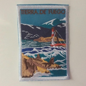 Puede incluir: Parche bordado con una escena costera y el texto "TIERRA DE FUEGO". El diseño incluye un faro, un velero y olas en tonos azules, blancos y marrones. El parche tiene un borde blanco.