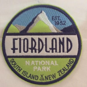 May include: Circular embroidered patch with a mountain range design. The patch features the text "FIORDLAND NATIONAL PARK SOUTH ISLAND NEW ZEALAND" and "EST. 1952". The colour scheme is blue, green, and white. Ideal for travel enthusiasts.