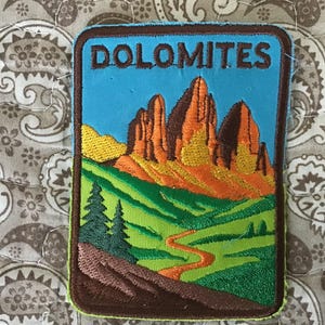 May include: A rectangular embroidered patch featuring the word DOLOMITES in brown letters at the top. The design depicts orange and brown mountain peaks above a green landscape with two dark green pine trees and a winding orange path.
