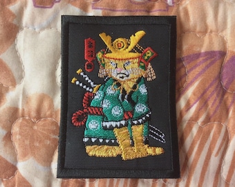Peut inclure: Écusson brodé représentant un chat samouraï de dessin animé. Le chat porte un kimono vert et blanc, des bottes jaunes et un casque jaune. Il a deux épées et une corde rouge. L'écusson est sur fond noir.