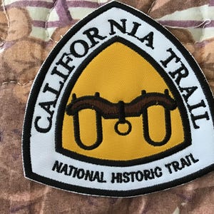 May include: A white, black, and yellow patch with the words "CALIFORNIA TRAIL" and "NATIONAL HISTORIC TRAIL" in black lettering. The patch features a yellow triangle with a brown design.
