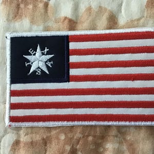 Peut inclure: Un écusson brodé rectangulaire arborant un motif inspiré du drapeau américain. Il présente un canton bleu foncé avec une étoile blanche et des symboles de boussole, suivis de bandes horizontales rouges et blanches avec une bordure blanche.