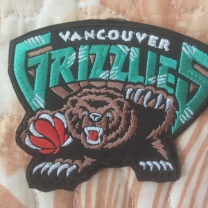 May include: Embroidered patch of the Vancouver Grizzlies logo. The patch features a black background with the team name in white and teal. A brown bear holding a basketball is the central image.
