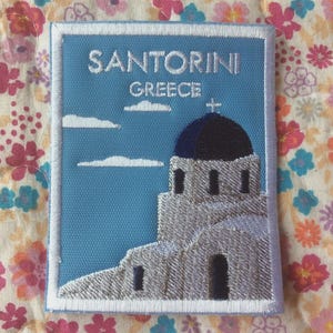 May include: Embroidered patch featuring a Santorini, Greece design. The patch has a blue background with white clouds and a church with a blue dome. The text "SANTORINI GREECE" is embroidered in white.