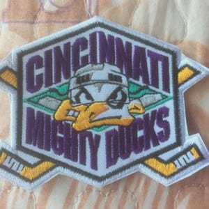 Könnte beinhalten: Gestickter Aufnäher mit dem Text "CINCINNATI MIGHTY DUCKS" in Lila. Der Aufnäher hat ein Hockey-Thema-Design mit einem Enten-Maskottchen, das einen Helm trägt. Der Aufnäher ist weiß mit gelben und türkisfarbenen Akzenten.