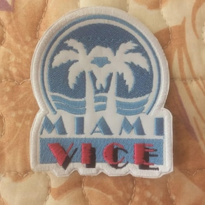 May include: Embroidered patch with a blue and white design of palm trees and the words "MIAMI VICE" in red and blue. The patch has a white border, suitable for application on clothing or accessories.