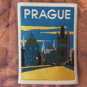 Op de afbeelding: Geborduurde patch met het woord "PRAGUE" boven een gedetailleerde afbeelding van de skyline van de stad. De patch heeft een blauwe achtergrond met gele accenten en een witte rand, met iconische architectuur.