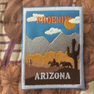 May include: Embroidered patch featuring a desert landscape with the text "PHOENIX ARIZONA". The design includes mountains, a sun, clouds, a saguaro cactus, and silhouettes of a horse and rider. The patch has a white border.