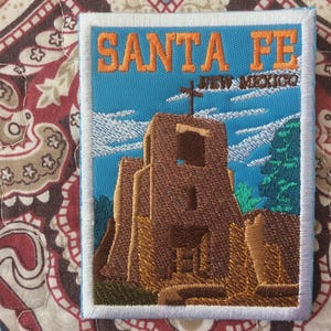May include: Embroidered patch depicting a Southwestern church with a cross, set against a blue sky. The patch reads "SANTA FE NEW MEXICO" in orange lettering. The patch has a white border and is on patterned fabric.
