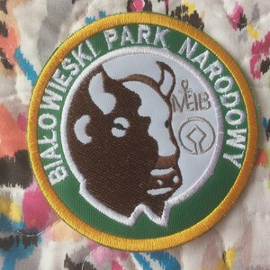May include: Embroidered circular patch with a bison head in brown and white, and the text "BIAŁOWIESKI PARK NARODOWY" in white on a green and gold border. The patch has a white background with additional text and symbols.