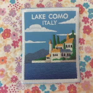 Pode incluir: Patch bordado com uma vista do Lago de Como, Itália. O design inclui um céu azul com nuvens brancas, um lago e edifícios com telhados laranja. O texto "LAKE COMO ITALY" está no topo.