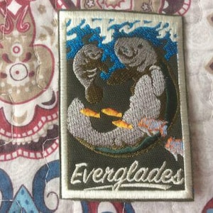 May include: Embroidered patch featuring two manatees in a marine scene with blue waves and orange and blue fish. The patch has a green background and the word "Everglades" in white script.