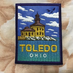 Könnte beinhalten: Gestickter Aufnäher mit einem gelben Gebäude mit braunem Dach und Turm, vor blauem Himmel mit weißen Wolken. Der Aufnäher trägt die Aufschrift "TOLEDO OHIO" in gelben und weißen Buchstaben, mit einem blauen Rand.