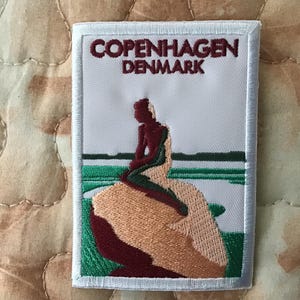 May include: A rectangular embroidered patch featuring a white background with the text COPENHAGEN DENMARK in dark red. The design depicts a stylized figure sitting on a large tan rock, surrounded by green and white water elements.