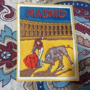May include: Embroidered patch featuring a bullfight scene in Madrid. The patch has a yellow background with a matador and bull. The word "MADRID" is embroidered in blue at the top. The patch is bordered in white and yellow.