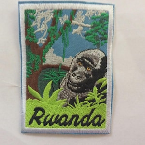 May include: Embroidered patch featuring a gorilla in a jungle setting with the word "Rwanda" in green and black. The patch has a light blue border and depicts a gorilla with yellow eyes.