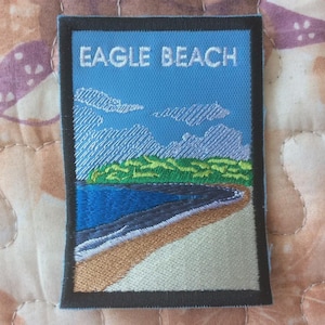 May include: Embroidered patch showing a beach scene with the text "EAGLE BEACH". The design includes a blue sky with clouds, a green treeline, and a blue ocean meeting a sandy shore. The patch has a black border.