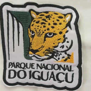 以下が含まれることがあります： 黄色と黒の斑点模様のジャガーの頭が刺繍されたパッチ。パッチには、黒い縁取りのある白い背景に「PARQUE NACIONAL DO IGUAÇU」という白文字が書かれています。