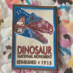 May include: Embroidered patch featuring a dinosaur skull over a blue and beige landscape. The patch reads "DINOSAUR NATIONAL MONUMENT ESTABLISHED 1915". The patch has a brown border and is rectangular.