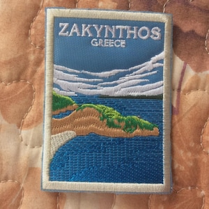 Könnte beinhalten: Gestickter Aufnäher mit einer Landschaft von Zakynthos, Griechenland. Das Design zeigt einen blauen Himmel, weiße Wolken und eine Küstenlinie mit grünem und braunem Land, das auf das blaue Meer trifft. Der Text "ZAKYNTHOS GREECE" ist weiß gestickt.