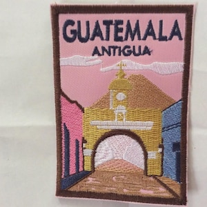 Puede incluir: Parche bordado que representa una escena de Guatemala, con el texto "GUATEMALA ANTIGUA". El parche muestra un arco amarillo y edificios en rosa y azul, sobre un cielo rosa con nubes blancas. Tiene un borde marrón.