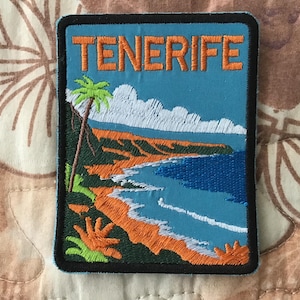 May include: Embroidered patch with the word "TENERIFE" in orange, depicting a coastal landscape. The design includes a palm tree, blue sea, white clouds, and orange cliffs, set against a light blue background with a black border.