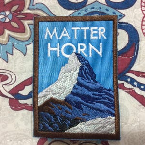 Può includere: Toppa ricamata raffigurante il Cervino, con la scritta "MATTER HORN" in bianco. La toppa ha uno sfondo blu e un bordo marrone. La montagna è raffigurata in tonalità di blu, bianco e marrone.
