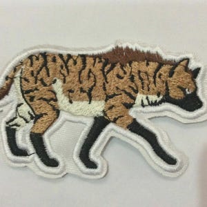 Puede incluir: Parche bordado con un diseño de hiena. El parche tiene una combinación de colores marrón y negro con un borde blanco. La hiena se representa en una pose de caminar, con sus patas y cabeza claramente definidas. El parche es probablemente para ropa o accesorios.