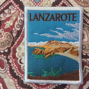 Pode incluir: Patch bordado com uma paisagem costeira com a palavra "LANZAROTE" em letras castanhas. O patch mostra uma costa com falésias castanhas, água verde e nuvens brancas. O patch retangular tem uma borda branca.