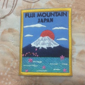 Puede incluir: Parche bordado con el monte Fuji en Japón. El diseño incluye un sol rojo, una montaña con cima nevada, cielo azul y agua con flores rosas. El parche tiene un borde amarillo y el texto "FUJI MOUNTAIN JAPAN".
