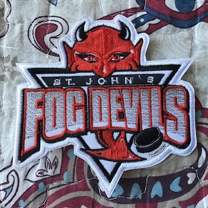 May include: An embroidered patch featuring the St. John's Fog Devils logo. The design includes a red devil head with black horns, white text reading ST. JOHN'S, large red and white lettering reading FOG DEVILS, and a black hockey puck graphic.