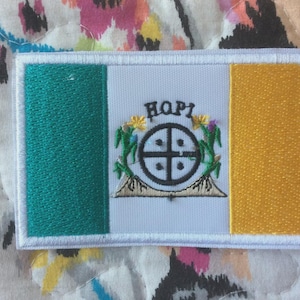 Puede incluir: Parche bordado con el diseño de la bandera Hopi. El parche tiene un fondo blanco con una franja vertical verde a la izquierda y una franja vertical amarilla a la derecha. El centro presenta un emblema negro con la palabra "HOPI" encima.